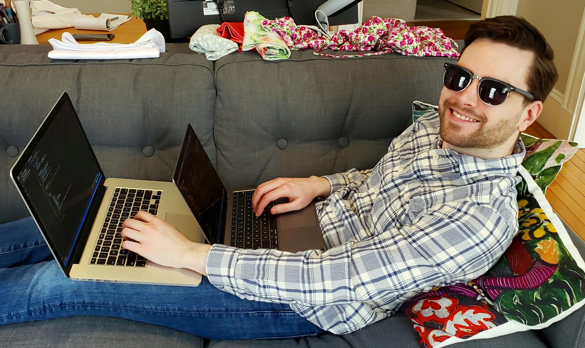 Andrew lying on a couch wearing sunglasses, somehow typing on two laptops simultaneously, smiling at the camera